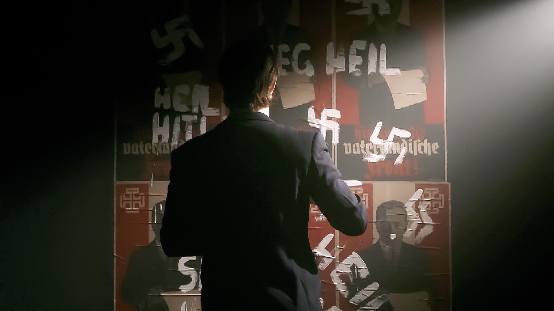 Das Bild zeigt eine dunkle Szene, in der eine Person in einem Anzug von hinten zu sehen ist. Die Person steht vor einer Wand, die mit mehreren Plakaten bedeckt ist. Die Plakate zeigen symbolische Elemente und Texte in roter und schwarzer Schrift, darunter ein Hakenkreuz und die Worte „Heil Hitler“. Einige der Texte sind mit schwarzer oder weißer Farbe überklebt oder durchgestrichen, was einen Akt der Zensur oder Ablehnung darstellt. Das Licht fällt von einer Quelle oben rechts, wodurch eine dramatische Licht- und Schattensituation entsteht und die Umgebung verstärkt wird. Die Atmosphäre ist ernst und historisch aufgeladen, was auf das Thema des Bildes hindeutet.
