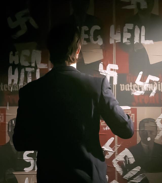 Das Bild zeigt eine dunkle Szene, in der eine Person in einem Anzug von hinten zu sehen ist. Die Person steht vor einer Wand, die mit mehreren Plakaten bedeckt ist. Die Plakate zeigen symbolische Elemente und Texte in roter und schwarzer Schrift, darunter ein Hakenkreuz und die Worte „Heil Hitler“. Einige der Texte sind mit schwarzer oder weißer Farbe überklebt oder durchgestrichen, was einen Akt der Zensur oder Ablehnung darstellt. Das Licht fällt von einer Quelle oben rechts, wodurch eine dramatische Licht- und Schattensituation entsteht und die Umgebung verstärkt wird. Die Atmosphäre ist ernst und historisch aufgeladen, was auf das Thema des Bildes hindeutet.