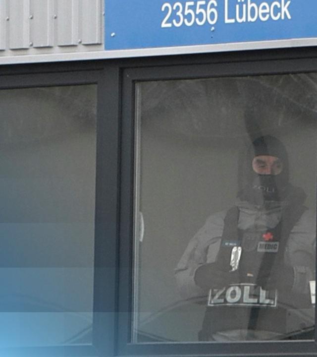 Zwei vermummte Zollbeamte stehen am Fenster eines Containers.