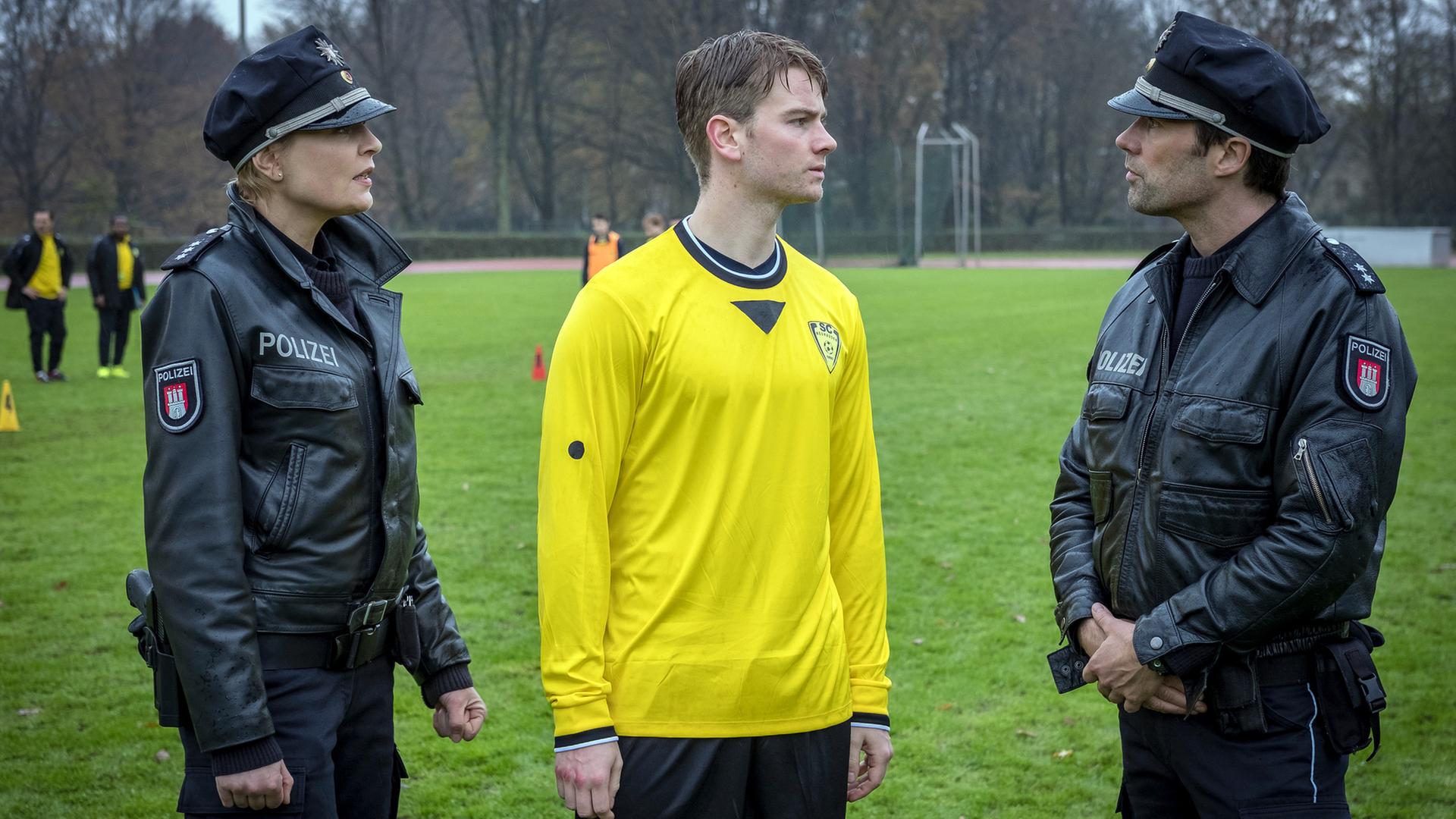 "Notruf Hafenkante - Im Abseits": Die Polizisten Melanie Hansen (Sanna Englund) und Mattes Seeler (Matthias Schloo) stehen auf einem Fußballplatz. Zwischen ihnen steht der Fußballer Yanik Jungbluth (Aaron Koszuta). Er trägt ein gelb-schwarzes Trikot und sieht Mattes an, der rechts neben ihm steht.
