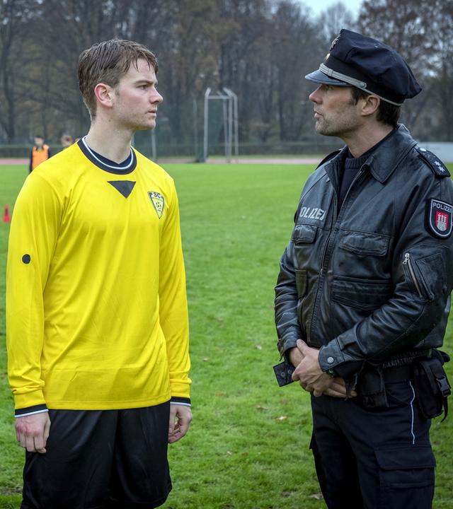 "Notruf Hafenkante - Im Abseits": Die Polizisten Melanie Hansen (Sanna Englund) und Mattes Seeler (Matthias Schloo) stehen auf einem Fußballplatz. Zwischen ihnen steht der Fußballer Yanik Jungbluth (Aaron Koszuta). Er trägt ein gelb-schwarzes Trikot und sieht Mattes an, der rechts neben ihm steht.