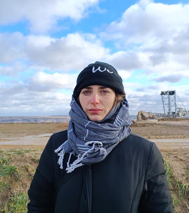 Junge Frau in Mütze und Schal gehüllt blickt ernst in die Kamera und steht vor dem weiten, leeren Gelände von Lützeratz. Im Hintergrund ein Braunkohlebagger zu erahnen, blauer Himmel mit zahlreichen Wolkenformationen.