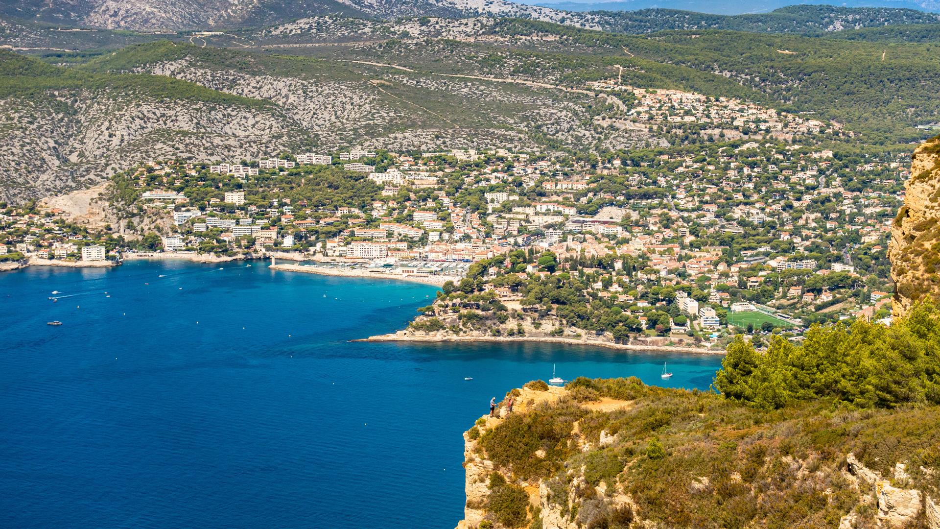 Blick auf die Bucht und die Altstadt von Cassis, eingerahmt von Felsen, Meer und Hügeln unter blauem Himmel.