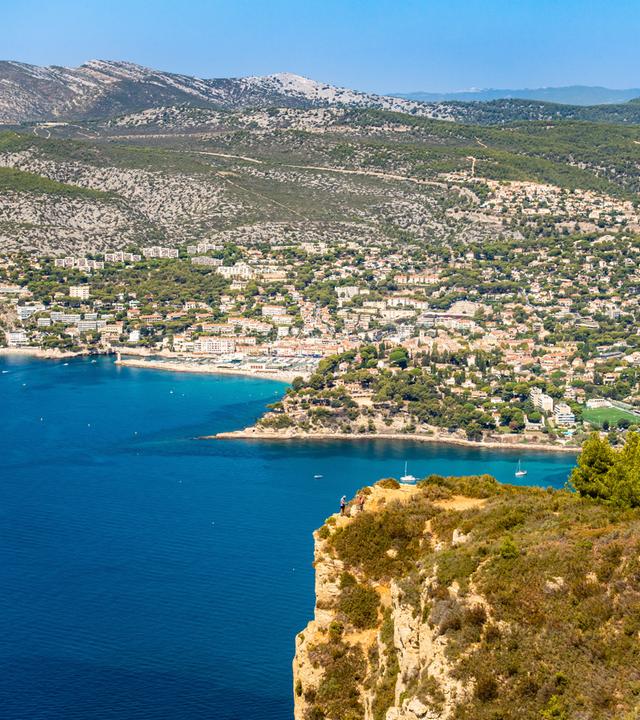Blick auf die Bucht und die Altstadt von Cassis, eingerahmt von Felsen, Meer und Hügeln unter blauem Himmel.