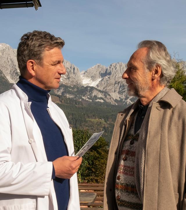 Vor der Arztpraxis stehen sich Dr. Martin Gruber (Hans Sigl) und Maurus Steiger (Daniel Friedrich) gegenüber. Lächelnd hält der Arzt ein Dokument in den Händen.