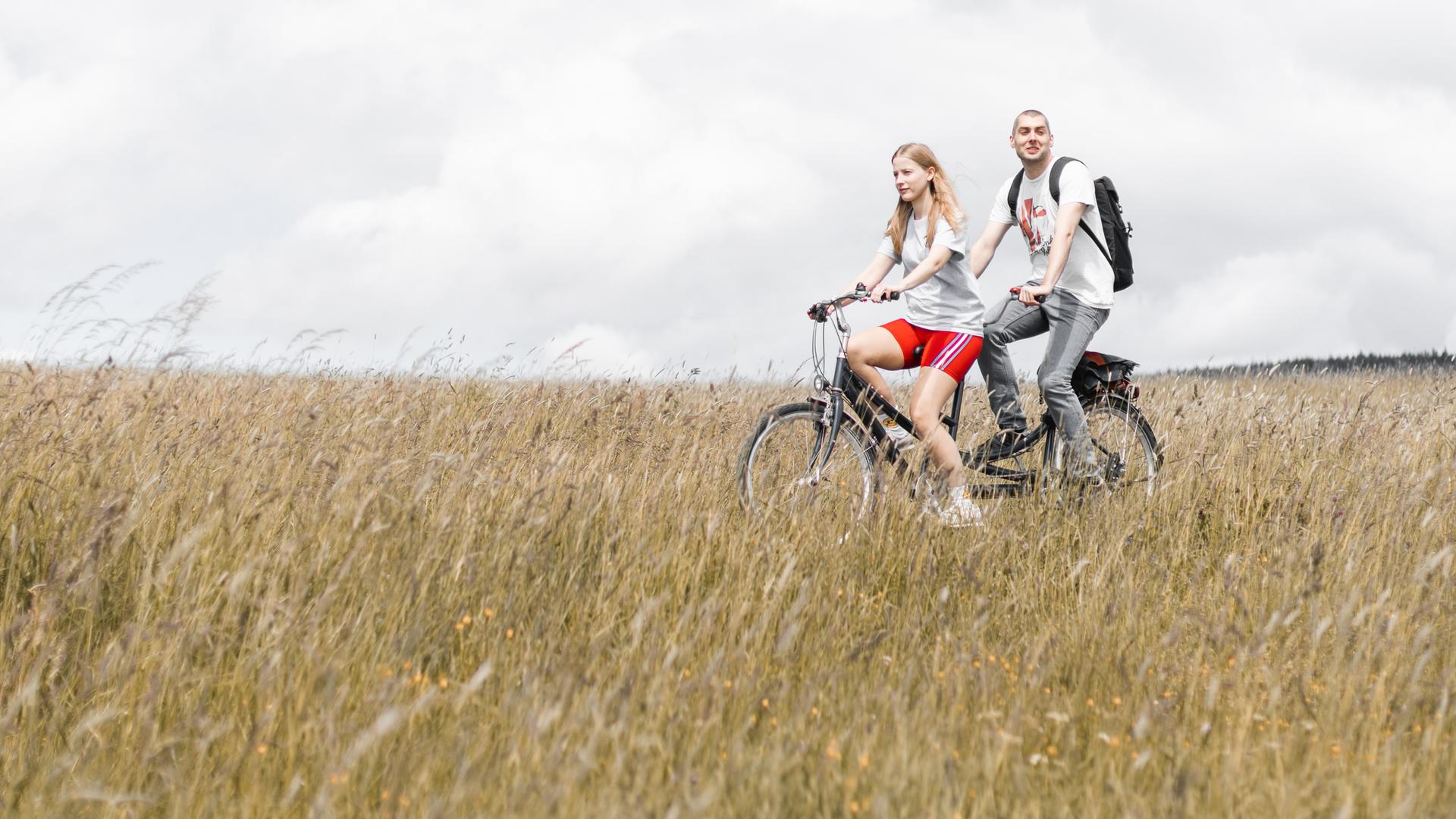 David und Cindy sitzen auf einem Tandem (Cindy vorn, David hinten) und fahren durch ein Feld, welches im Vordergrund zu sehen ist. Im Hintergrund sieht man einen bewölkten Himmel.