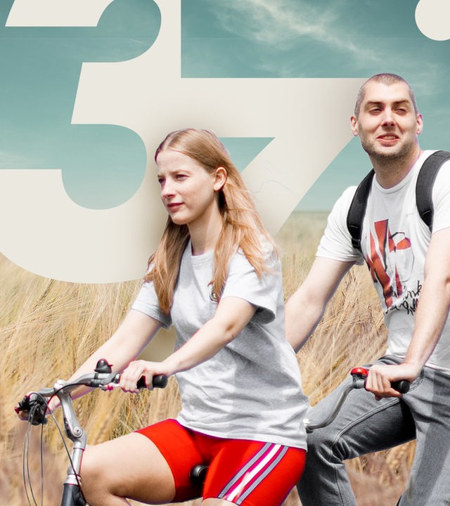 David und Cindy sitzen auf einem Tandem (Cindy vorn, David hinten) und fahren durch ein Feld, welches im Vordergrund zu sehen ist. Im Hintergrund sieht man einen bewölkten Himmel und ein Kornfeld.