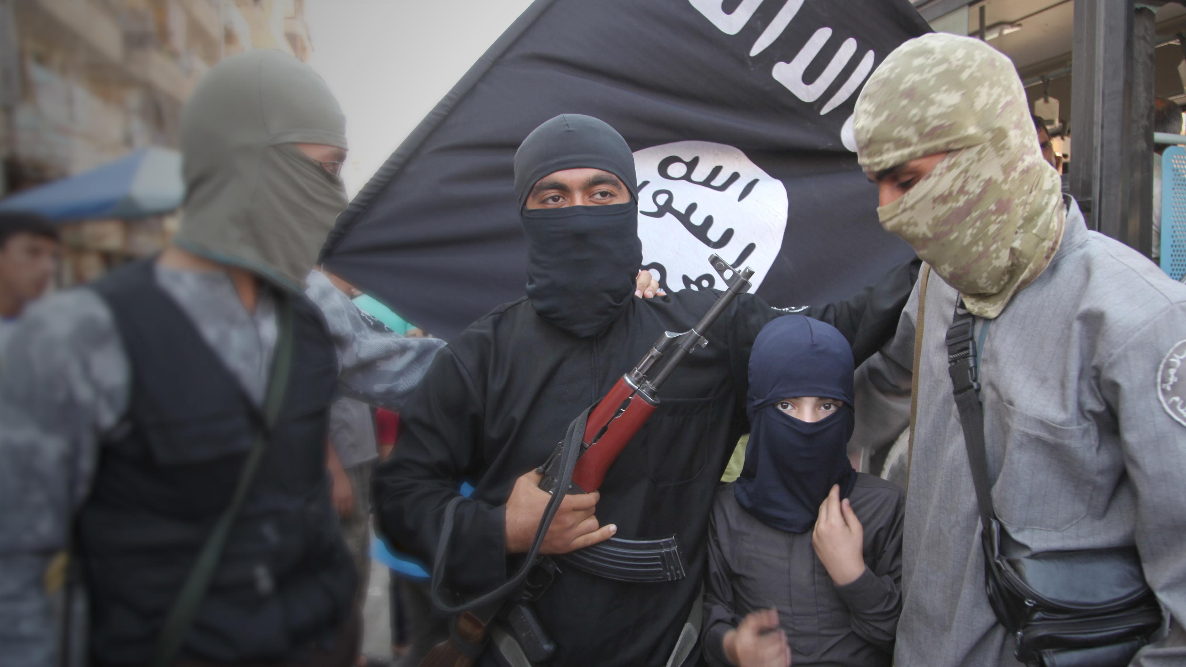 Eine Gruppe IS-Soldaten, darunter ein Kind, posieren mit Waffen vor ihrer Flagge.