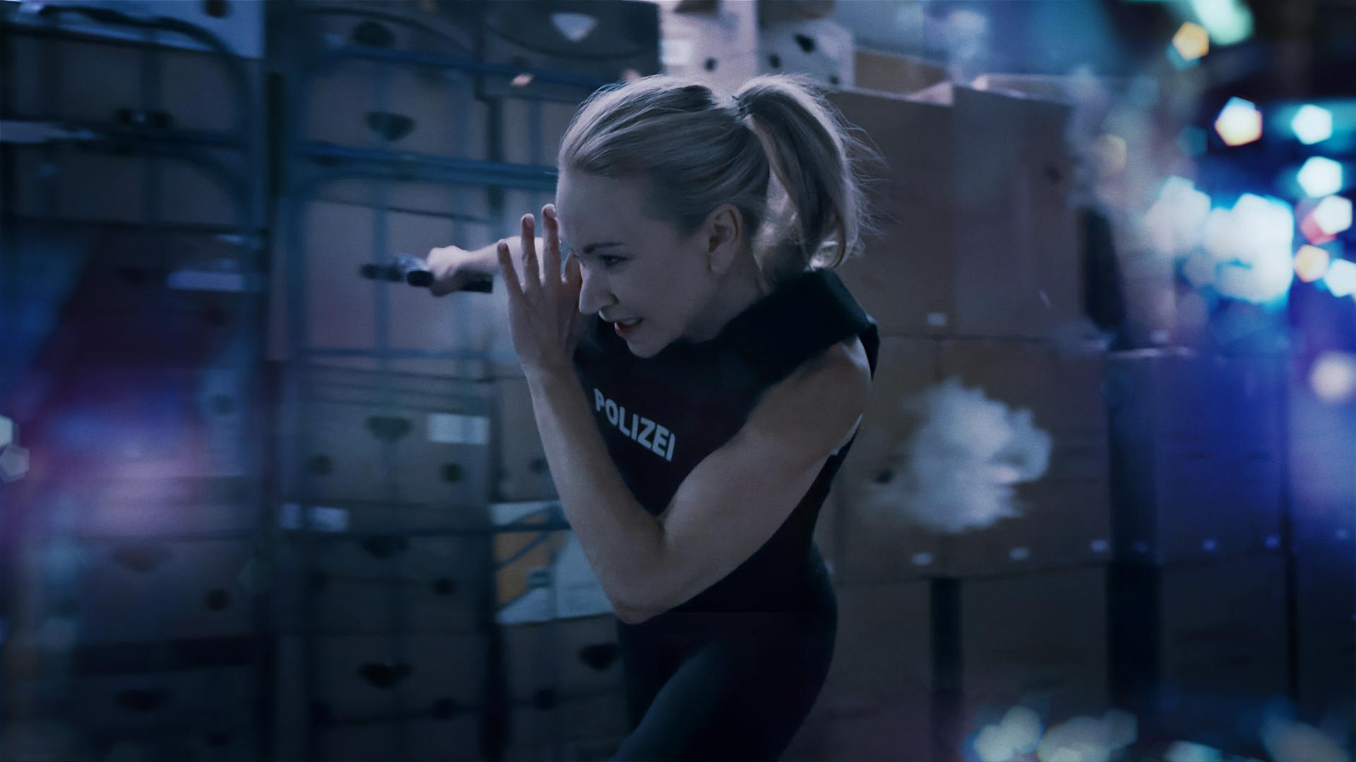 Sarah Kohr (Lisa Maria Potthoff), eine sportliche blonde Frau in Polizeiweste, rennt durch ein Warenlager. Schüsse schlagen in die Pakete hinter ihr ein. In der einen Hand hält sie eine Waffe.
