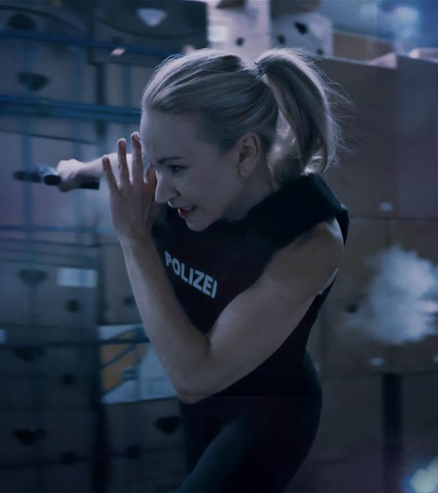 Sarah Kohr (Lisa Maria Potthoff), eine sportliche blonde Frau in Polizeiweste, rennt durch ein Warenlager. Schüsse schlagen in die Pakete hinter ihr ein. In der einen Hand hält sie eine Waffe.