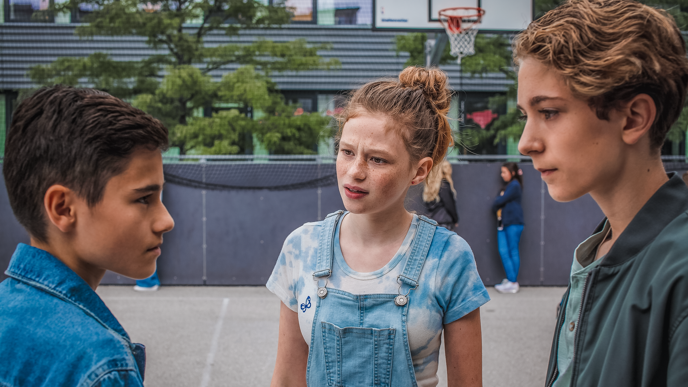 Charly steht zwischen Benny und Hamid und schaut Hamid vorwurfsvoll an. Die beiden Jungs schauen sich ernst in die Augen.