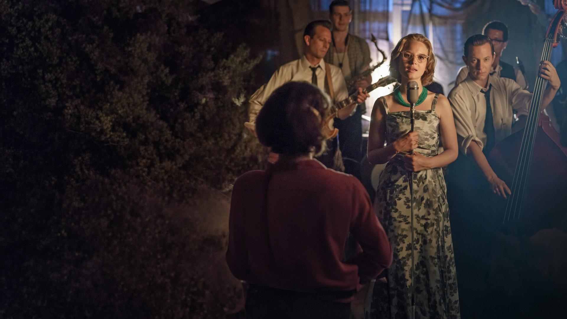 "Ku'damm 59 - 3. Im Urwald": Monika Schöllack (Sonja Gerhardt) steht auf einer Bühne und singt, während Freddy Donath (Trystan Pütter) etwas hinter ihr Kontrabass spielt. Weitere Musiker stehen hinter den beiden.