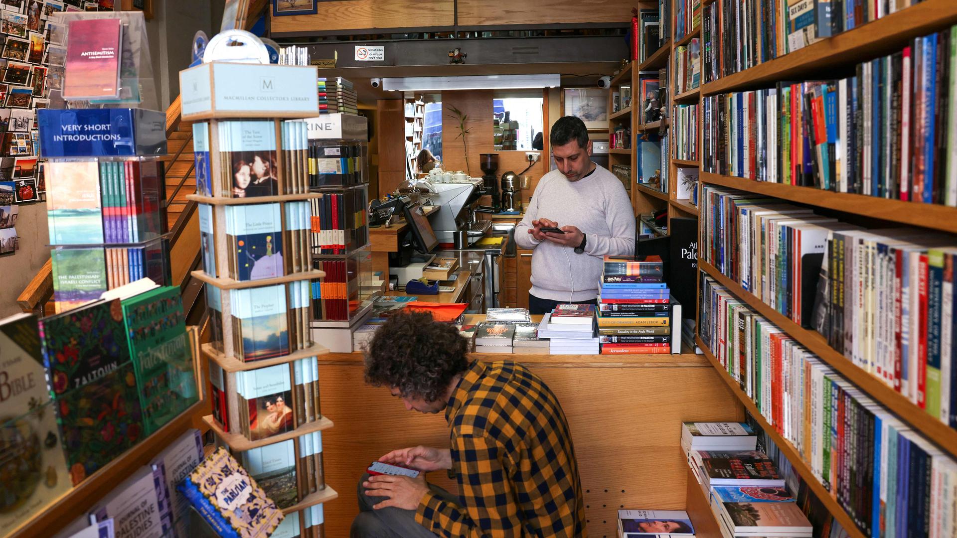 In einer Buchhandlung sitzt ein Mann im Vordergrund und schaut auf sein Handy, hinter ihm steht ein Mann an der Kasse und schaut auf sein Handy.