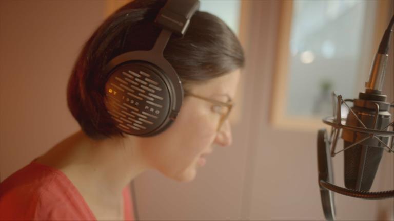 Nahaufnahme einer Sprecherin mit Kopfhörern, die in ein Studiomikrofon spricht. Der Screenshot veranschaulicht, wie Audiodeskription für ZDF-Inhalte aufgenommen wird.