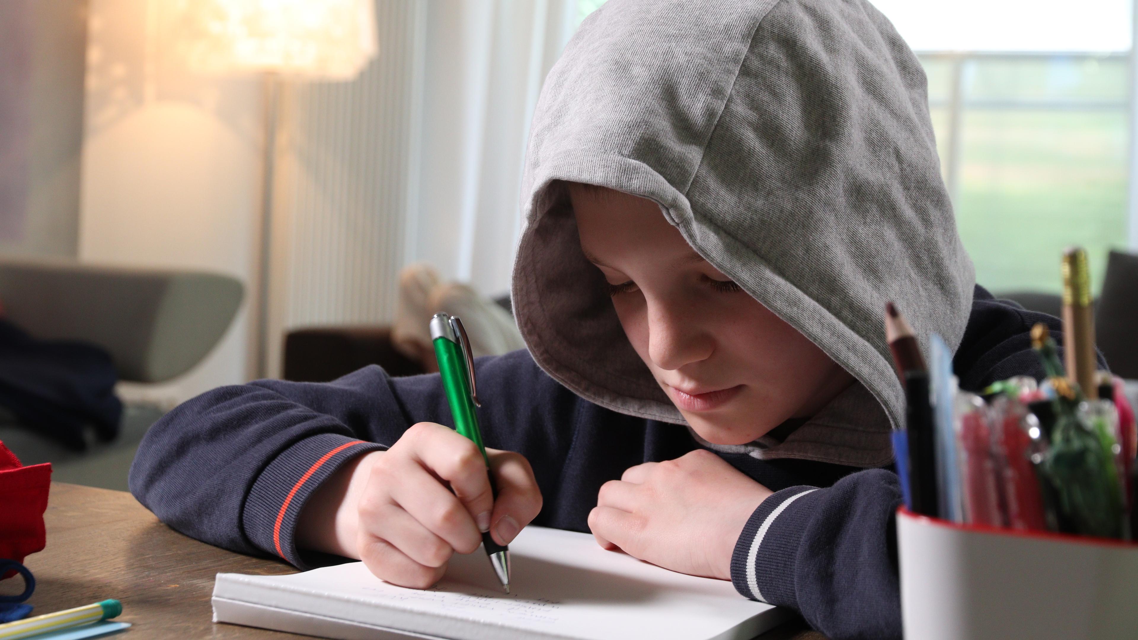 Wenn das Alphabet zur Qual wird": Bela Lamm sitzt am Tisch und hat eine Kapuze auf. Vor ihm liegt ein Papierblock. In seiner rechten Hand hält er einen Kugelschreiber.