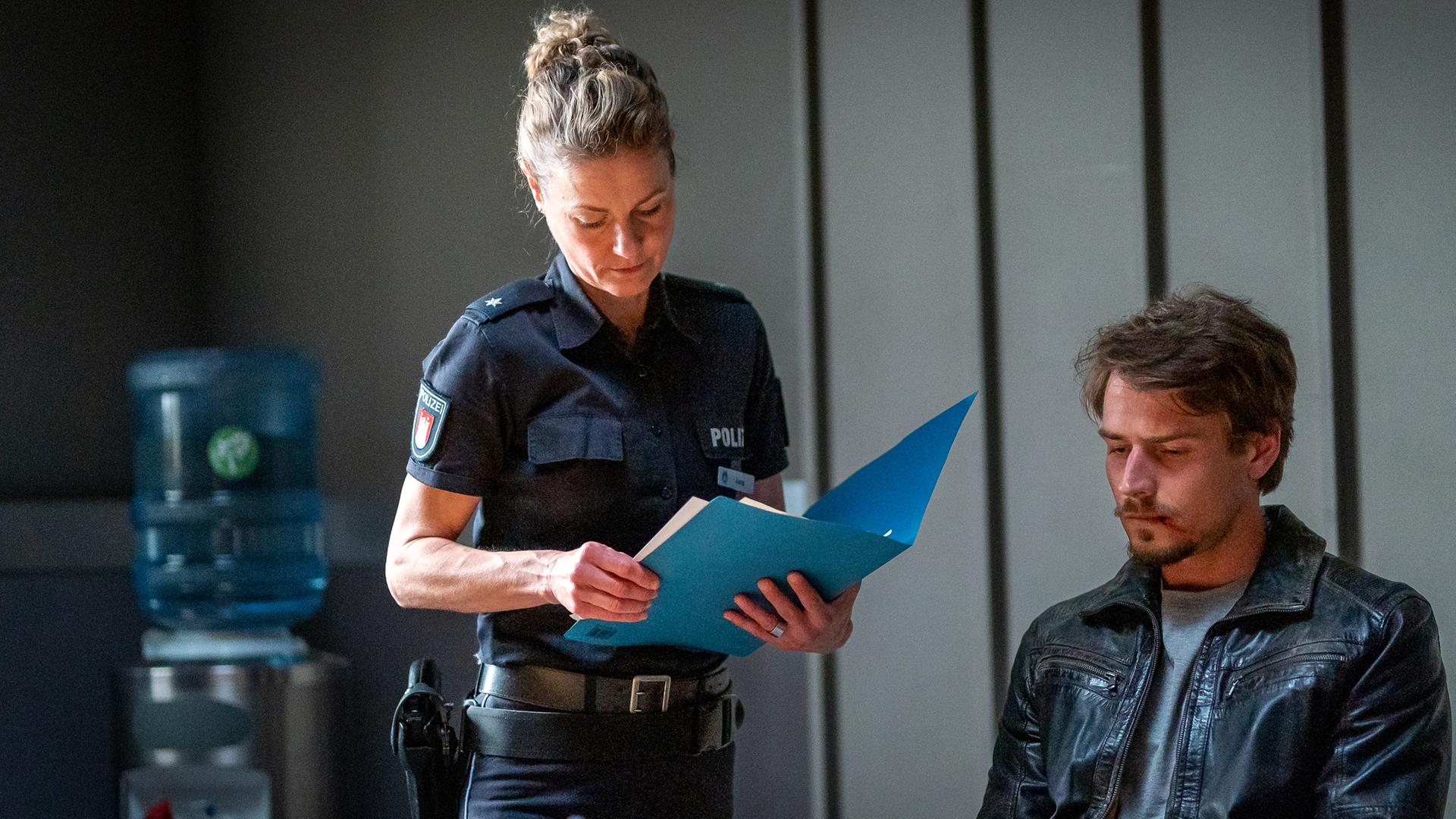 Steffen Bremer (Ric Wersig) sitzt an einem Tisch im Befragungsraum. Er blickt mürrisch auf ein Foto, das vor ihm auf dem Tisch liegt. Die Polizistin Franzi Jung (Rhea Harder-Vennewald) steht neben ihm. Sie hält eine Akte in den Händen und blickt auf die Papiere, die darin liegen.