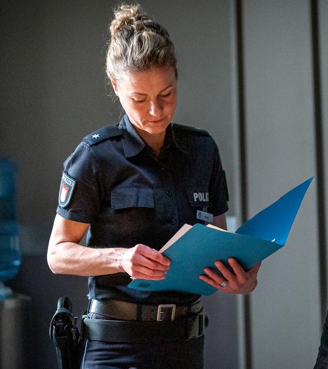 Steffen Bremer (Ric Wersig) sitzt an einem Tisch im Befragungsraum. Er blickt mürrisch auf ein Foto, das vor ihm auf dem Tisch liegt. Die Polizistin Franzi Jung (Rhea Harder-Vennewald) steht neben ihm. Sie hält eine Akte in den Händen und blickt auf die Papiere, die darin liegen.