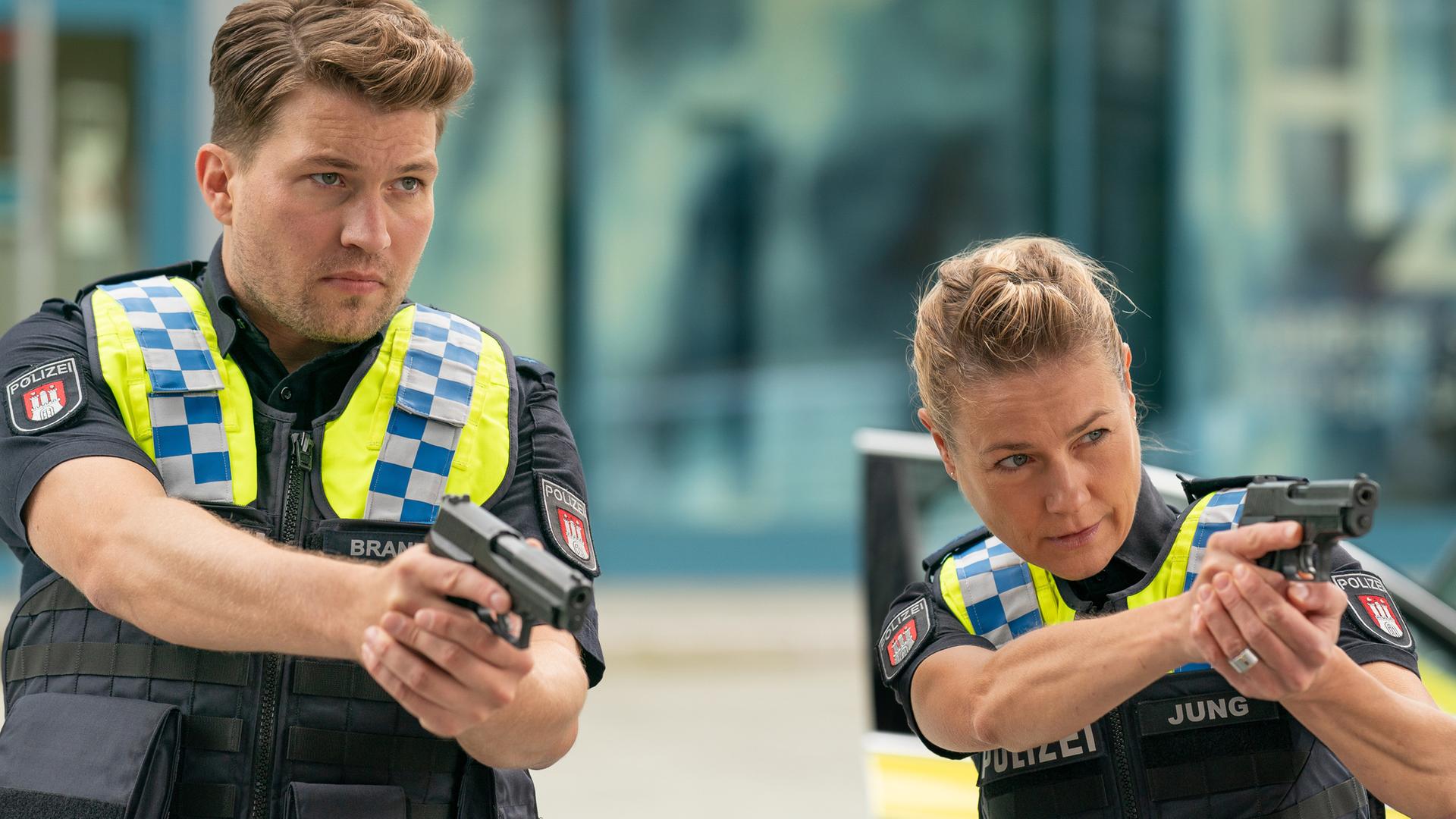 Der Polizist Nick Brandt (Raúl Richter) und seine Kollegin Franzi Jung (Rhea Harder-Vennewald) stehen nebeneinander. Beide zielen mit ihrer Waffe in die gleiche Richtung.