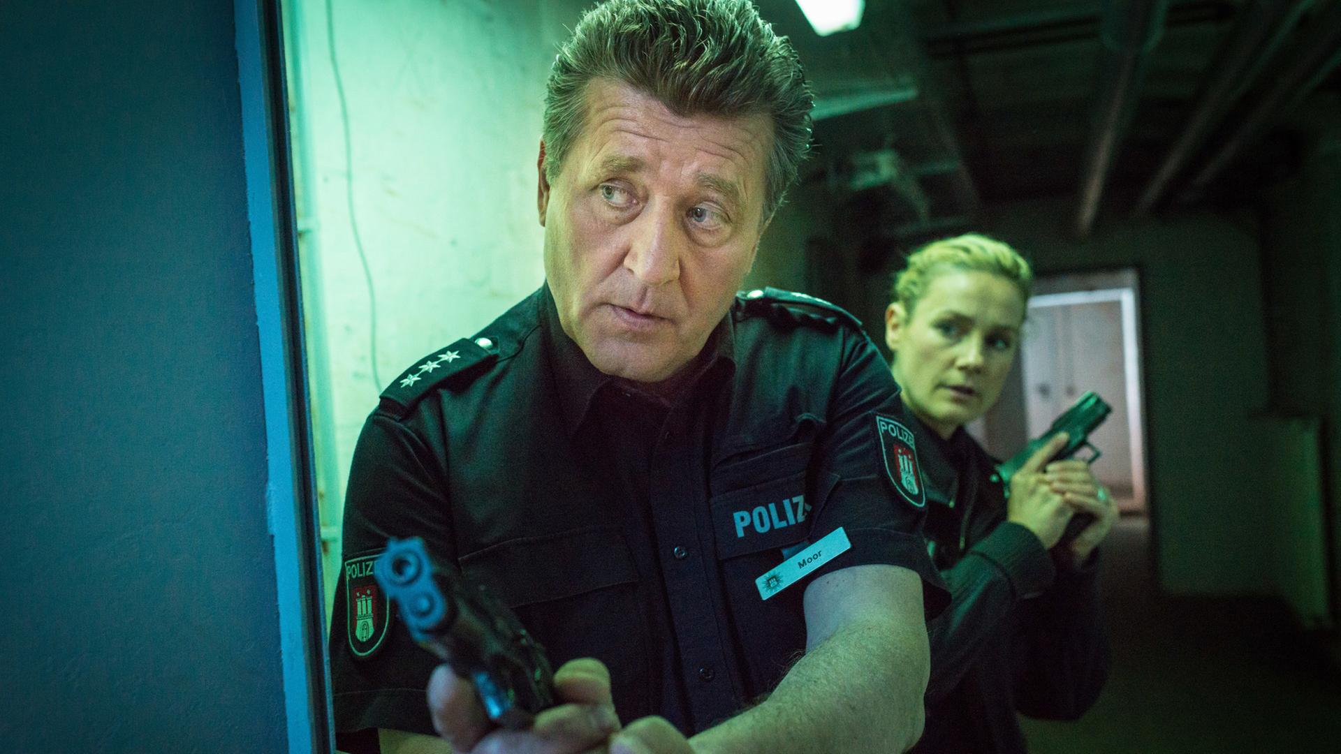 "Notruf Hafenkante - In Hamburg essen sie Hunde": In einem düsteren Kellergang stehen die Polizeibeamten Hans Moor (Bruno F. Apitz) und Franzi Jung (Rhea Harder-Vennewald) mit gezogenen Waffen hintereinander in Deckung.