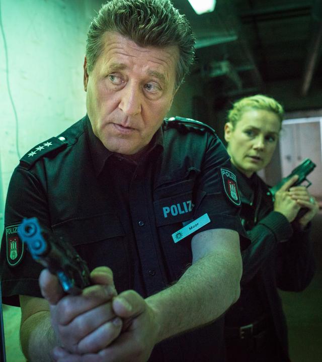 "Notruf Hafenkante - In Hamburg essen sie Hunde": In einem düsteren Kellergang stehen die Polizeibeamten Hans Moor (Bruno F. Apitz) und Franzi Jung (Rhea Harder-Vennewald) mit gezogenen Waffen hintereinander in Deckung.