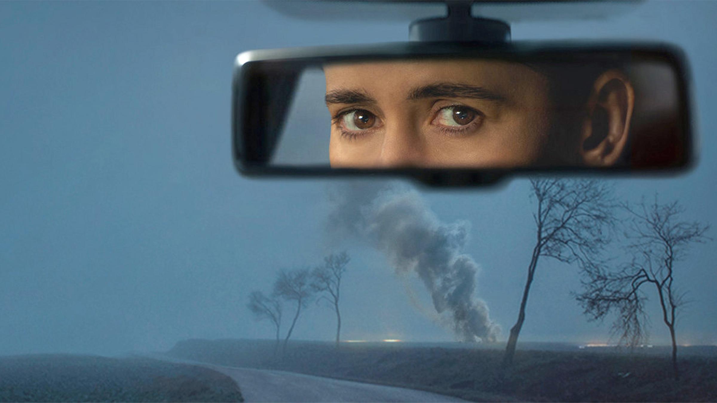 "In Her Car" (Keyvisual): Blick durch die Windschutzscheibe eines Autos in der Dämmerung. Am Horiziont steigen Rauchschwaden auf. Im Innenrückspiegel das Gesicht der Fahrerin.