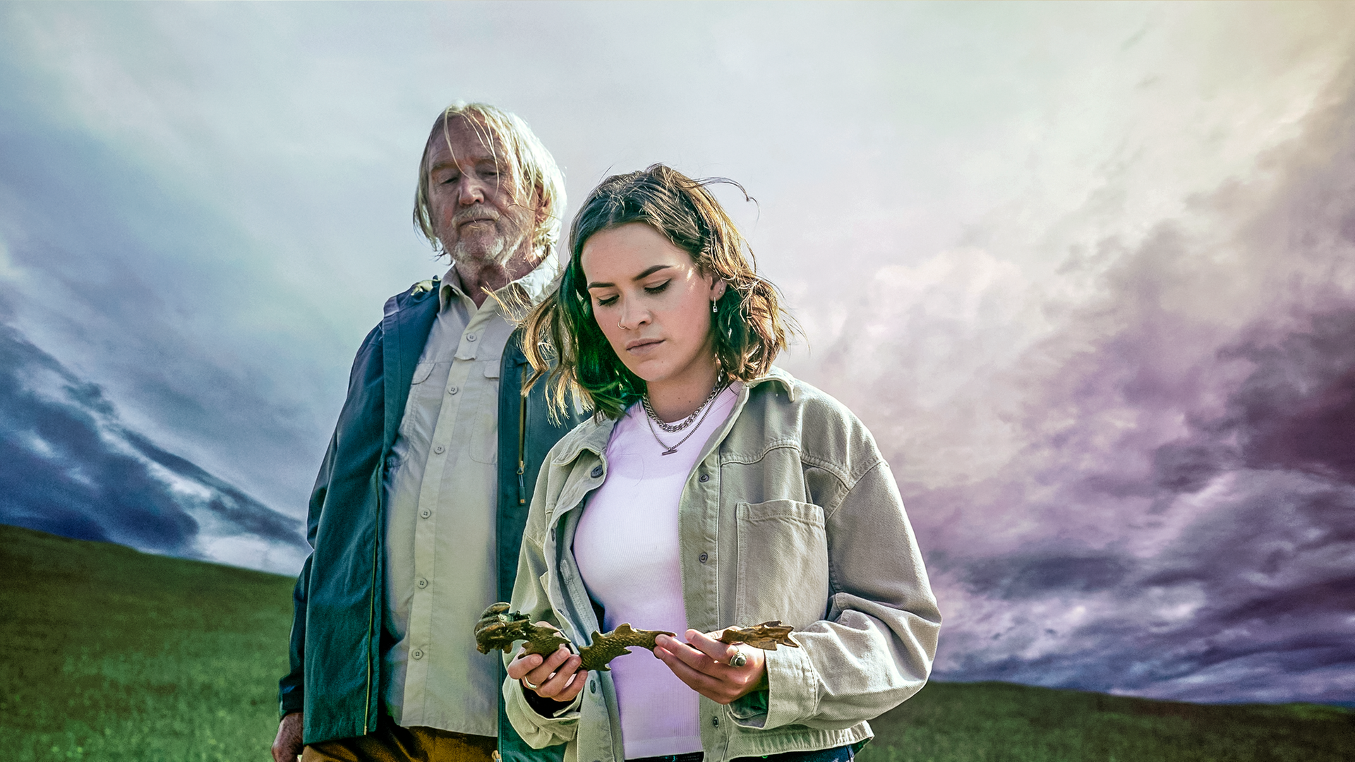 Issie (rechts) steht neben ihrem Großvater Gordon (links), der mit starren Blick auf die goldene Drachenfigur schaut, die Issie in ihren Händen hält. Um sie herum beginnt ein Unwetter aufzuziehen.
