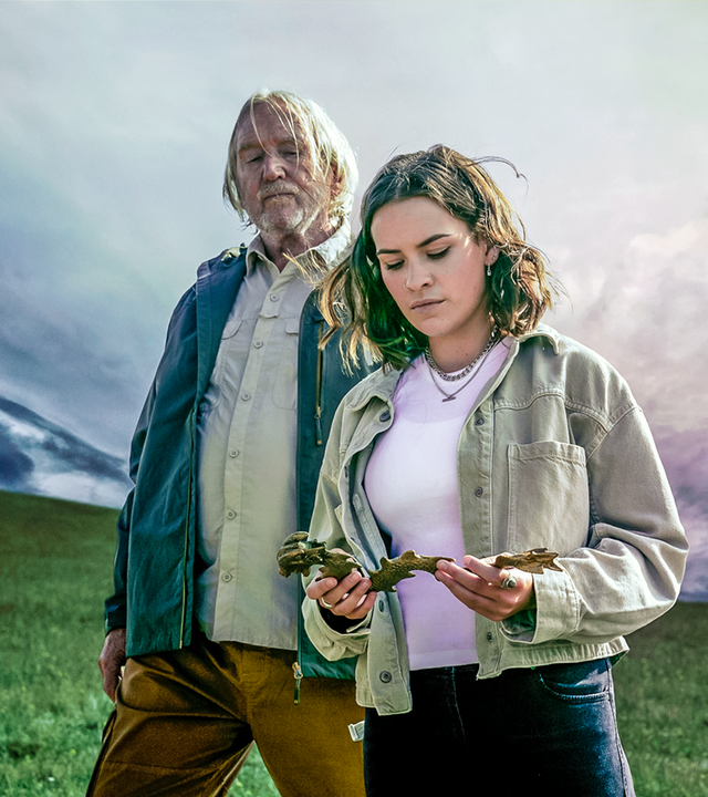 Issie (rechts) steht neben ihrem Großvater Gordon (links), der mit starren Blick auf die goldene Drachenfigur schaut, die Issie in ihren Händen hält. Um sie herum beginnt ein Unwetter aufzuziehen.