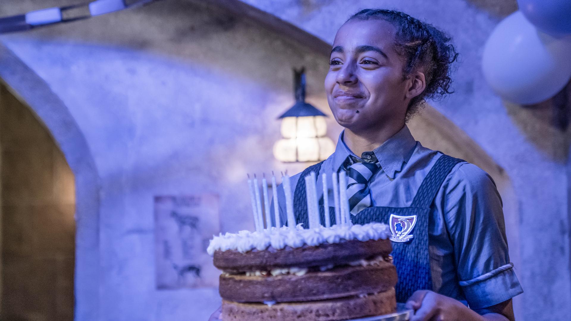 Indigo hat Geburtstag! Sie steht in einem Zimmer mit Blaulicht und hält glücklich eine große, runde, dreistöckige Torte mit bereits ausgeblasenen Kerzen vor sich in den Händen.