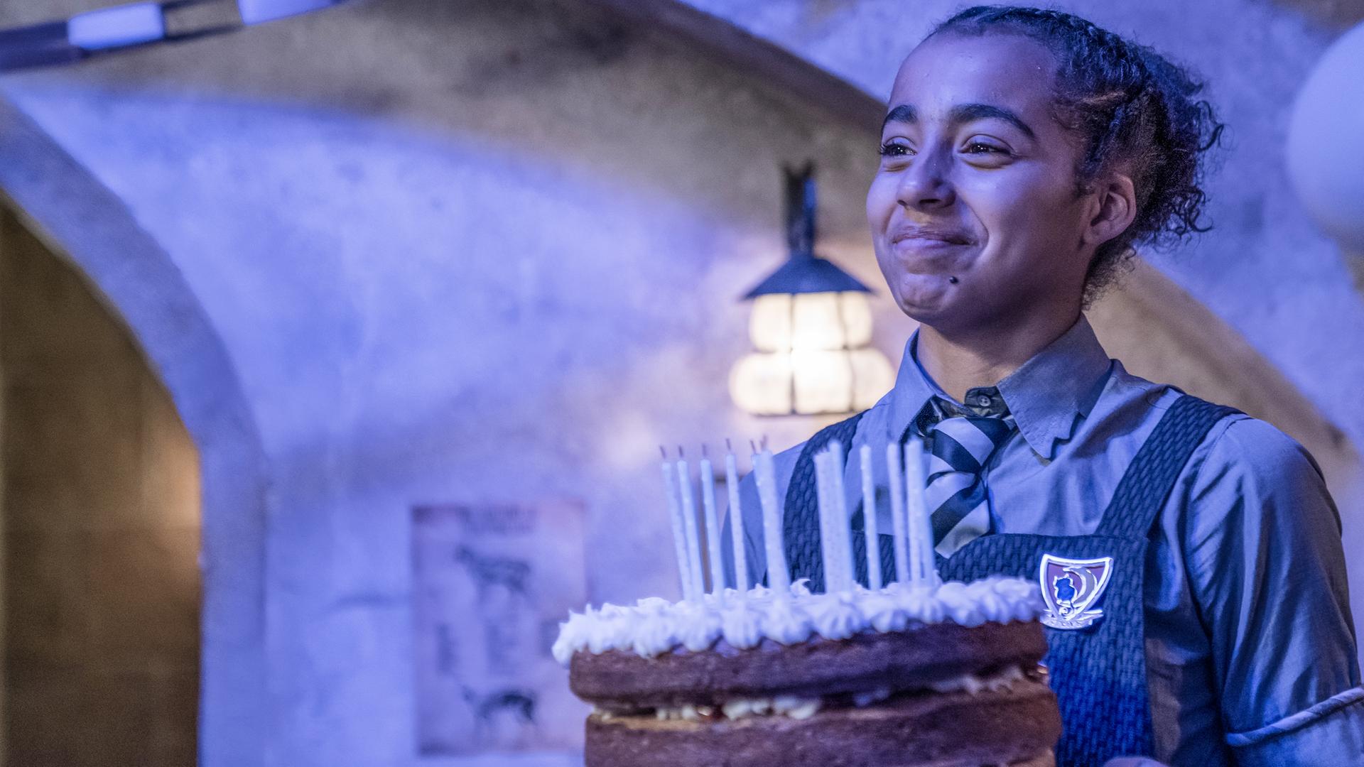 Indigo hat Geburtstag! Sie steht in einem Zimmer mit Blaulicht und hält glücklich eine große, runde, dreistöckige Torte mit bereits ausgeblasenen Kerzen vor sich in den Händen.