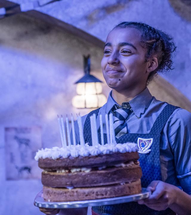 Indigo hat Geburtstag! Sie steht in einem Zimmer mit Blaulicht und hält glücklich eine große, runde, dreistöckige Torte mit bereits ausgeblasenen Kerzen vor sich in den Händen.