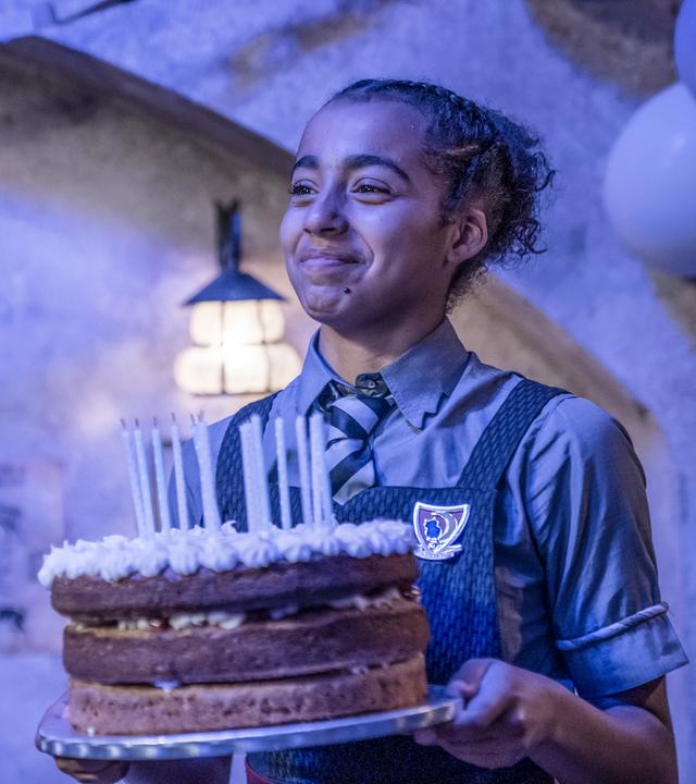 Indigo hat Geburtstag! Sie steht in einem Zimmer mit Blaulicht und hält glücklich eine große, runde, dreistöckige Torte mit bereits ausgeblasenen Kerzen vor sich in den Händen.
