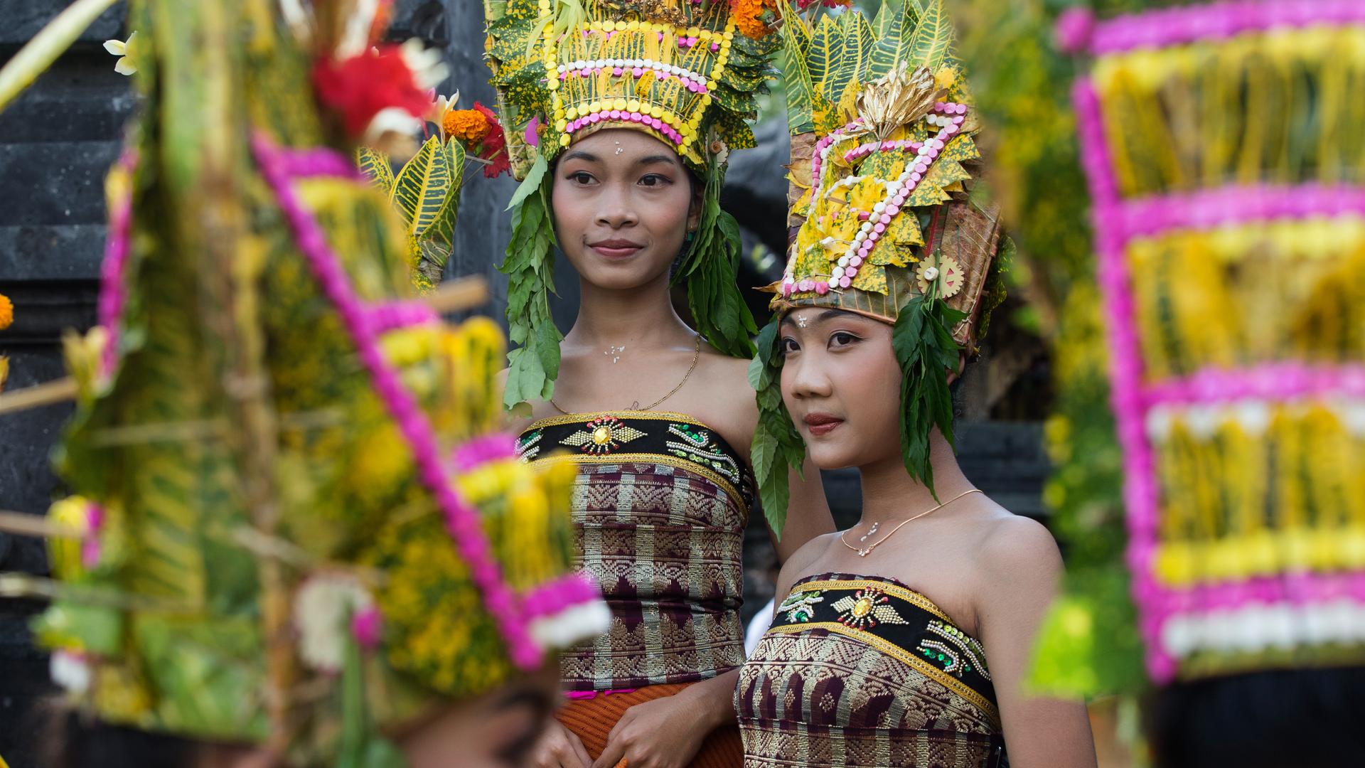 Zwei junge asiatisch aussehende Frauen tragen traditionellen balinesischen Kopfschmuck aus Blumen und Blättern und bestickte schulterfreie Wickelkleider.
