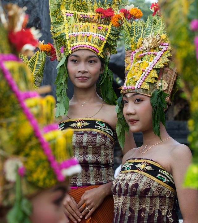 Zwei junge asiatisch aussehende Frauen tragen traditionellen balinesischen Kopfschmuck aus Blumen und Blättern und bestickte schulterfreie Wickelkleider.