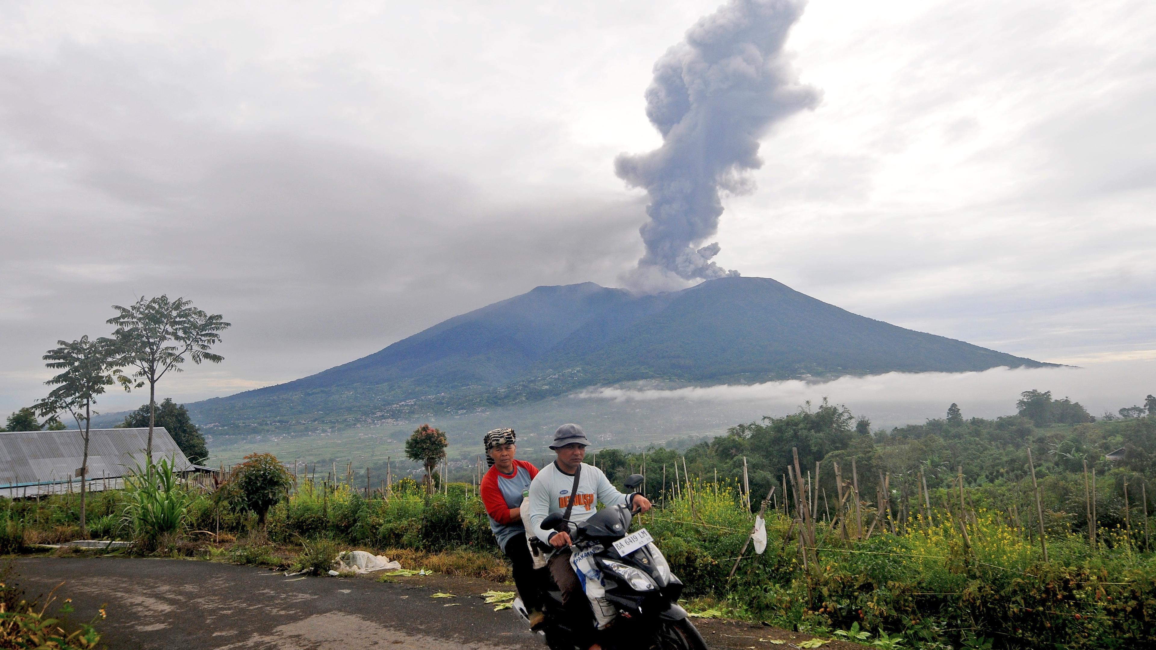Zwei Menschen fahren auf einem Mofa weg, im Hintergrund sieht man den Vulkan Marapi mit einer großen Rauchwolke.