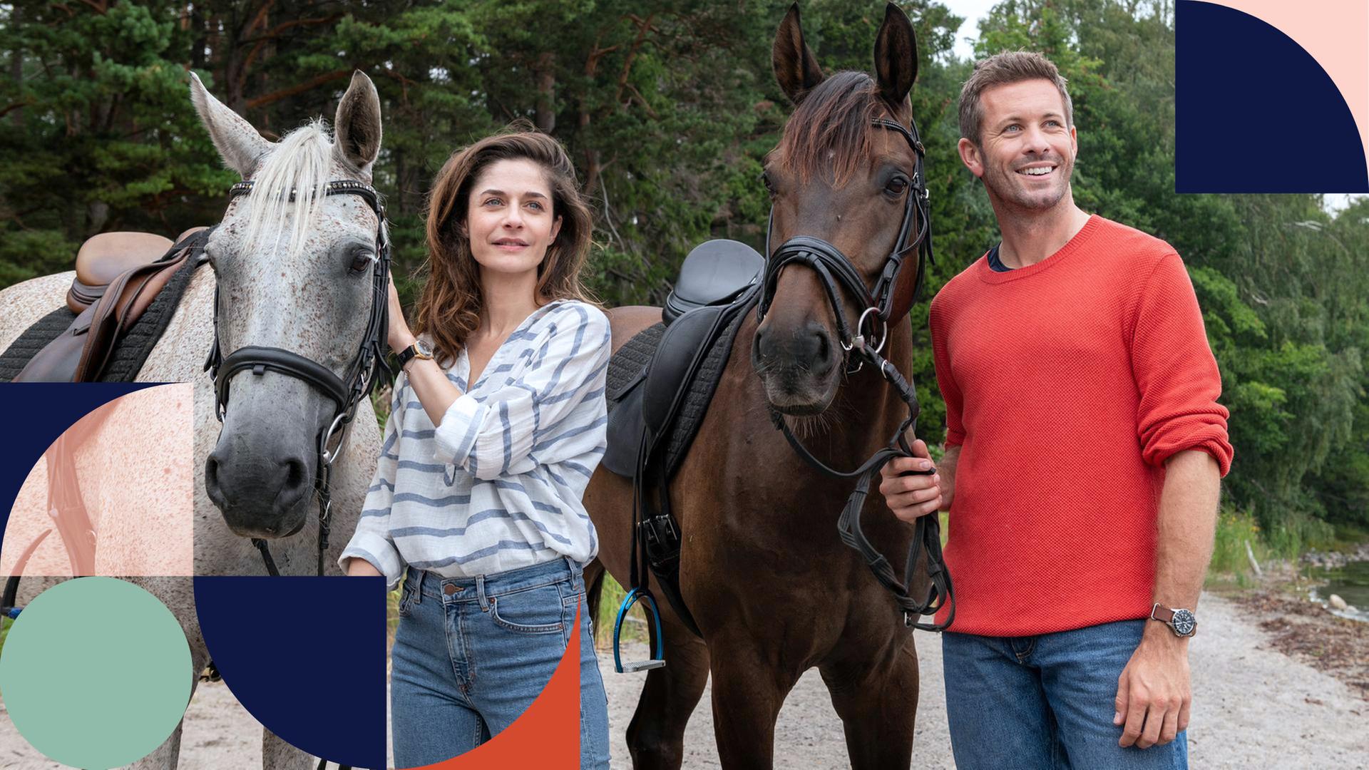 Eine Frau und ein Mann stehen beide neben einem weißen und einem braunen Pferd und schauen lächelnd in die Ferne.