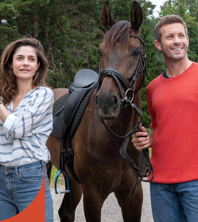 Emilia (Anne Werner) und Alexander (Jan Hartmann) stehen vor einem Wald neben ihren Pferden und schauen in die Ferne.
