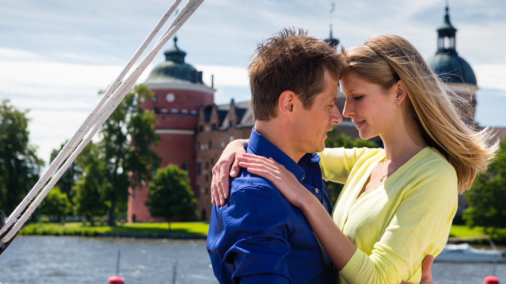 Leonie (Sarah Ulrich) und Erik (Roman Knižka) stehen eng umschlungen auf einem Boot und schauen sich dabei verliebt an. Hinter den beiden erstreckt sich ein Fluss. Auf der anderen Flussseite steht ein Schloss.
