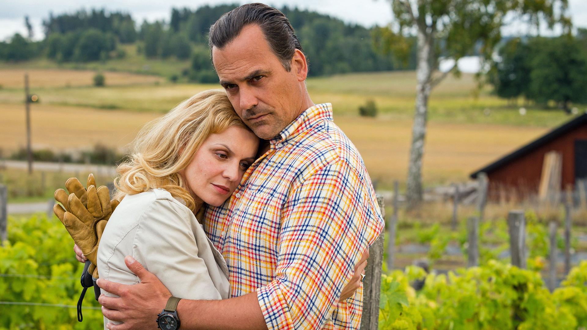 "Inga Lindström: Der Traum vom Siljansee": Elin Holmberg (Anja Nejarri) lehnt sich an Magnus Olofson (Marcus Grüsser), der sie umarmt. Beide schauen ernst und etwas besorgt. Im Hintergrund erstreckt sich ein weites Feld.