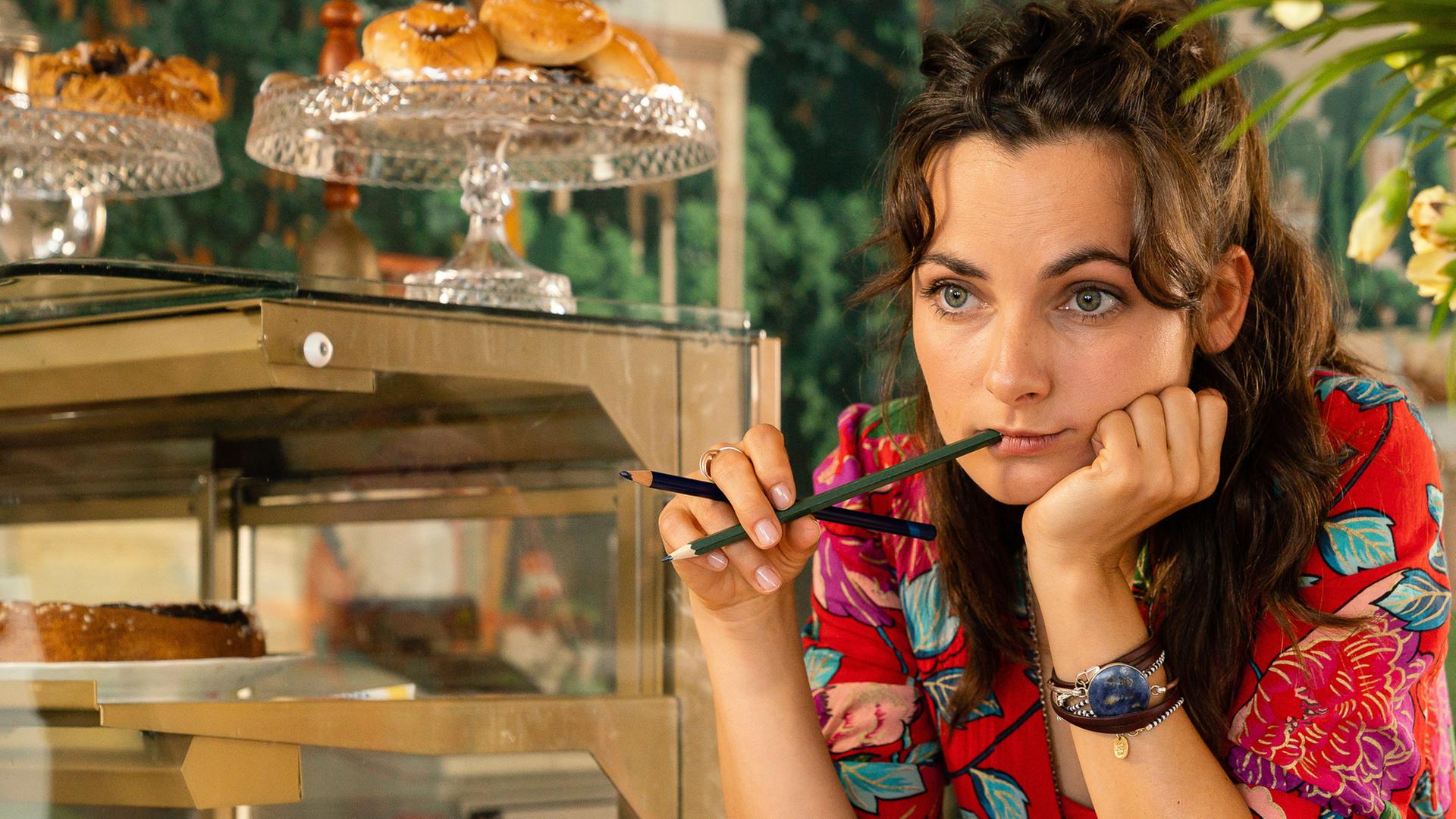 Jette (Paula Schramm), mit beiden Ellenbogen auf dem Tisch im Cafè abgestützt und einen Bleistift am Mund, träumt vor sich hin.