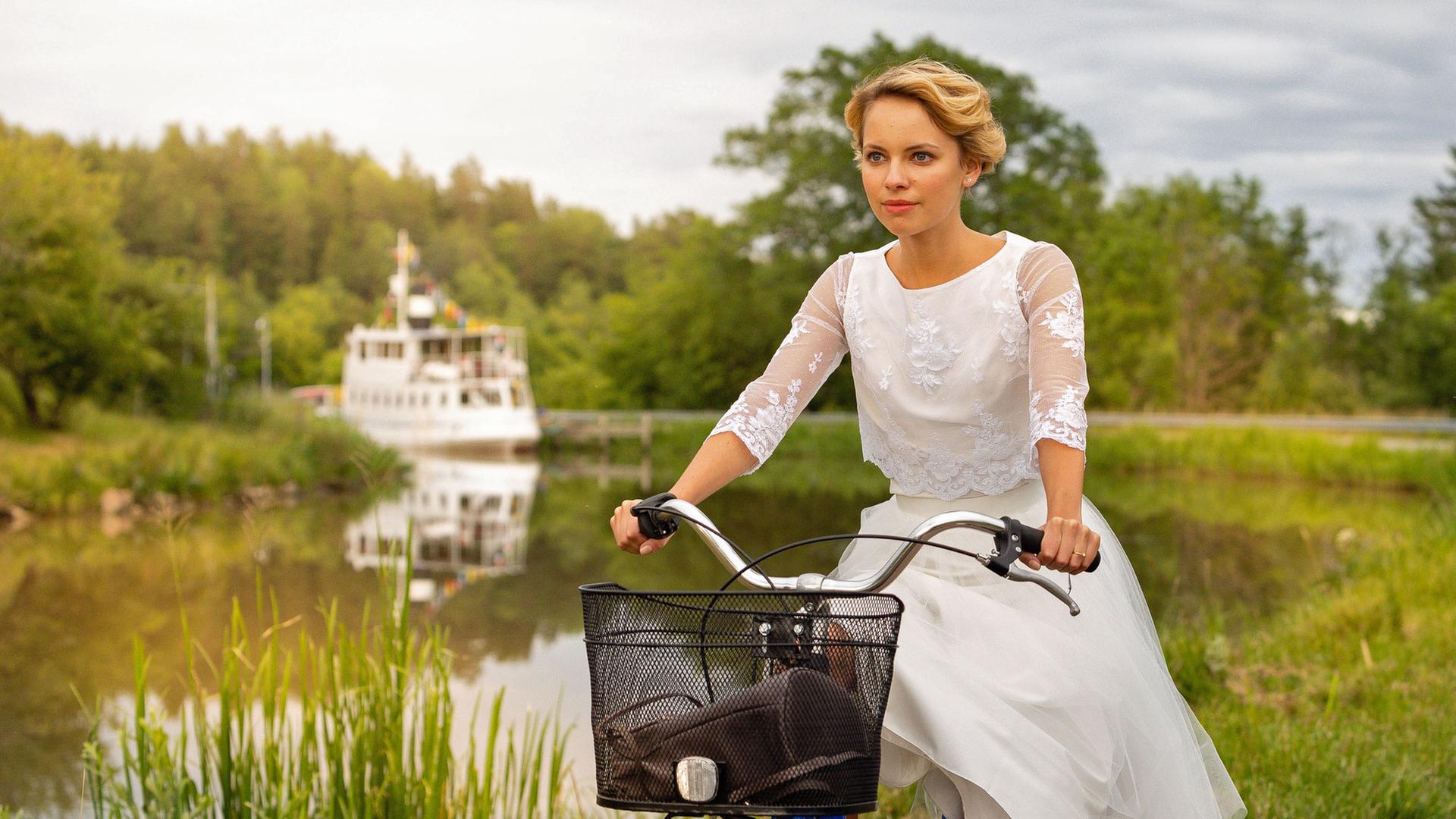 "Inga Lindström: Die Braut vom Götakanal": Emma (Barbara Prakopenka) fährt in einem weißen Kleid auf einem Fahrrad auf einem Feldweg im Grünen entlang des Götakanals. Im Hintergrund ist ein Schiff zu sehen.