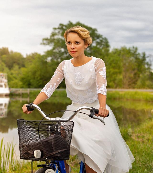 "Inga Lindström: Die Braut vom Götakanal": Emma (Barbara Prakopenka) fährt in einem weißen Kleid auf einem Fahrrad auf einem Feldweg im Grünen entlang des Götakanals. Im Hintergrund ist ein Schiff zu sehen.