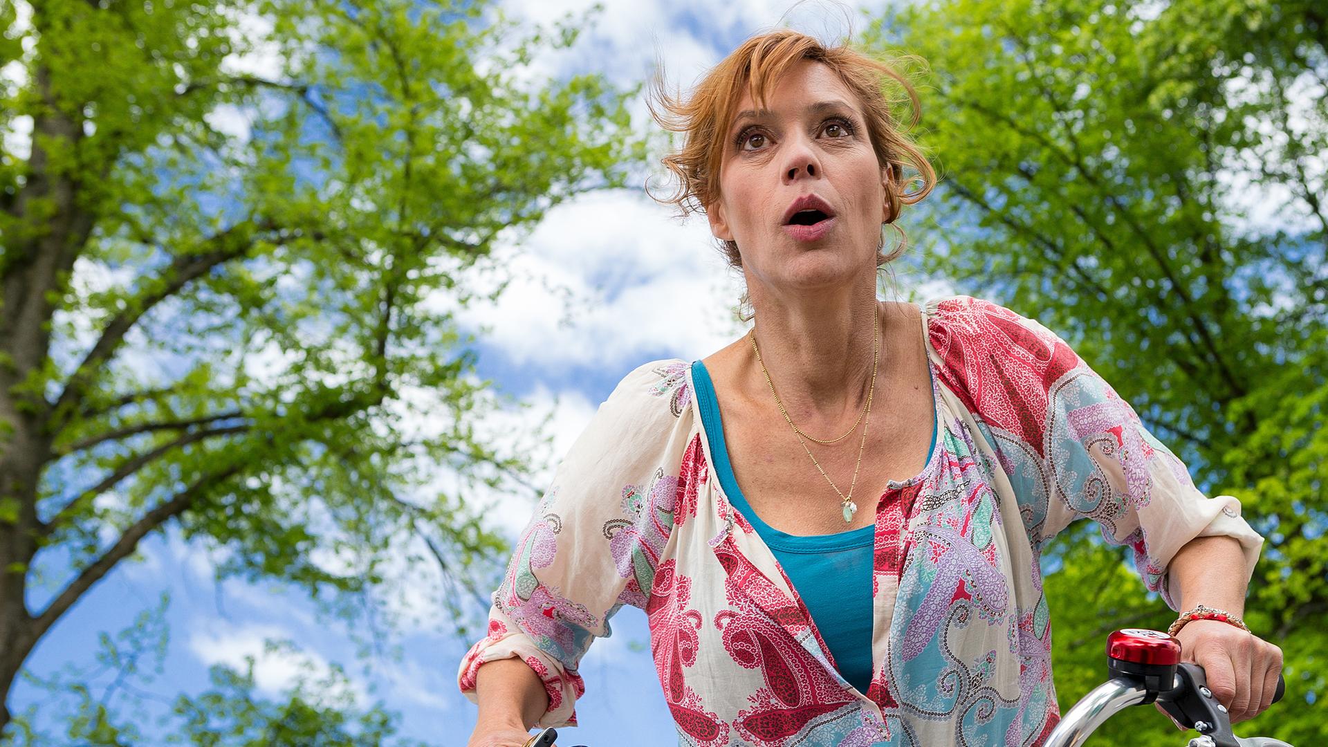 Unter bewölktem Himmel und Bäumen ist Eva Sellgren (Carin C. Tietze) mit dem Fahrrad unterwegs und schaut erschrocken.