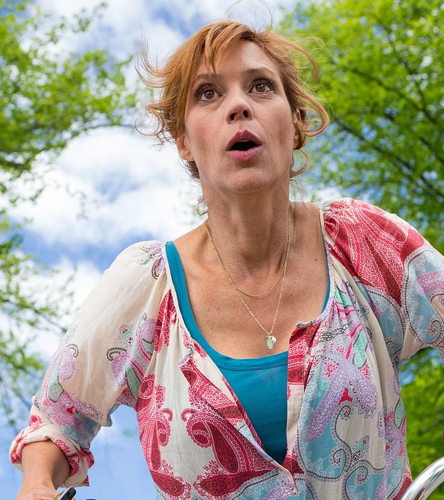 Unter bewölktem Himmel und Bäumen ist Eva Sellgren (Carin C. Tietze) mit dem Fahrrad unterwegs und schaut erschrocken.
