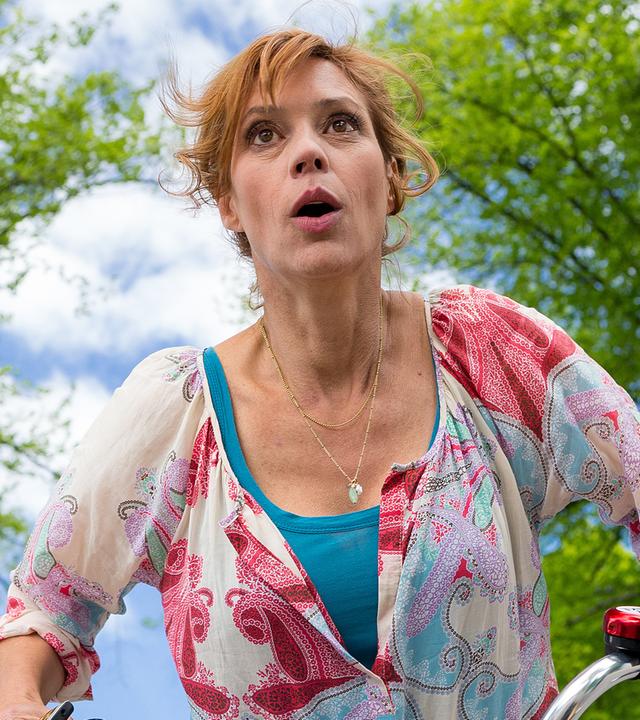 Unter bewölktem Himmel und Bäumen ist Eva Sellgren (Carin C. Tietze) mit dem Fahrrad unterwegs und schaut erschrocken.