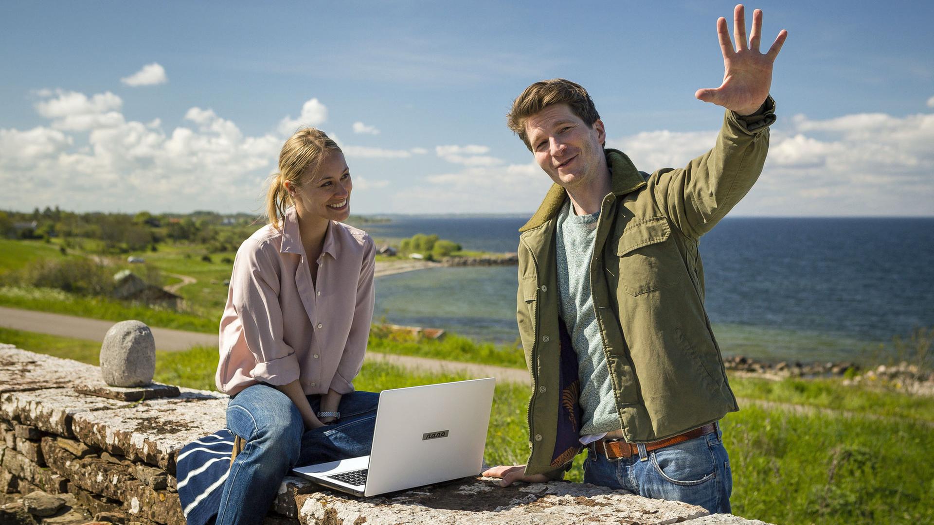 "Inga Lindström: Leg dich nicht mit Lilli an": Lilli Hellberg (Sina Tkotsch) sitzt mit Laptop auf einer Mauer, Malte Sommerfeld (Leopold Hornung) steht daneben, im Hintergrund ist das Meer zu sehen.