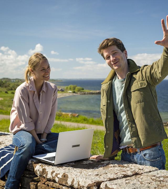 "Inga Lindström: Leg dich nicht mit Lilli an": Lilli Hellberg (Sina Tkotsch) sitzt mit Laptop auf einer Mauer, Malte Sommerfeld (Leopold Hornung) steht daneben, im Hintergrund ist das Meer zu sehen.