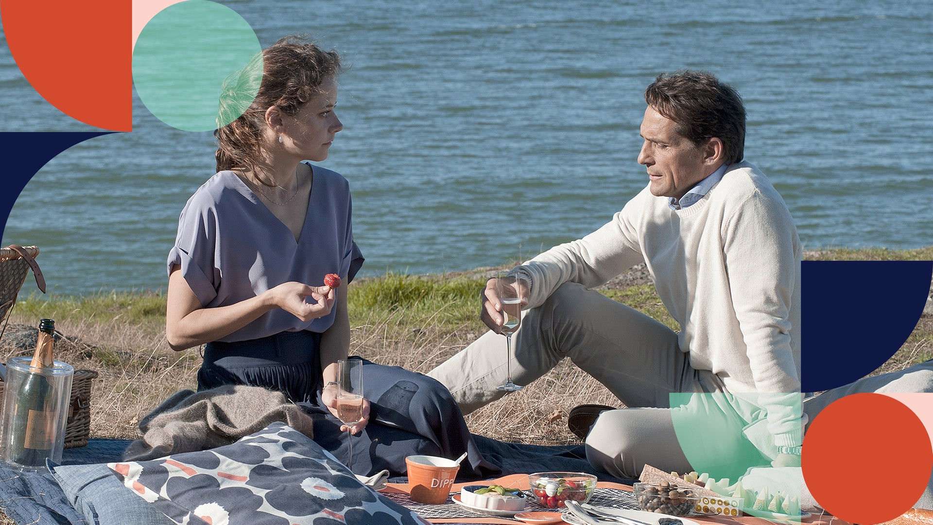 Sylvie und Lars sitzen auf einer Picknickdecke am See. Auf der Decke ist ein Picknick aufbereitet. Sylvie hält eine Kirsche, Lars ein Glas Sekt in der Hand. Sie schauen sich einander in die Augen.