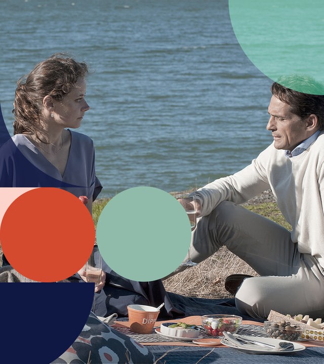 Sylvie und Lars sitzen auf einer Picknickdecke am See. Auf der Decke ist ein Picknick aufbereitet. Sylvie hält eine Kirsche, Lars ein Glas Sekt in der Hand. Sie schauen sich einander in die Augen.