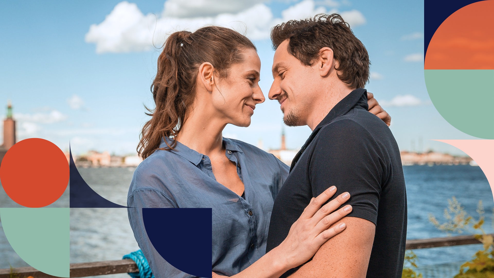 "Inga Lindström: Verliebt in meinen Chef": Anna Lund (Ellenie Salvo Gonzalez) und Sven Berggren (Markus Brandl) umarmen sich und schauen sich dabei an. Im Hintergrund: Wasser, Häuser und Stockholmer Stadtlandschaft.
