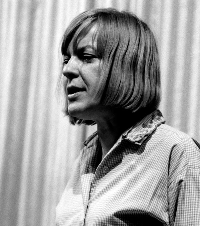 Altes Schwarz-Weiß-Foto einer Frau mit kinnlangen mittelblonden Haaren, die an einem Pult steht und etwas vorliest, oder redet.